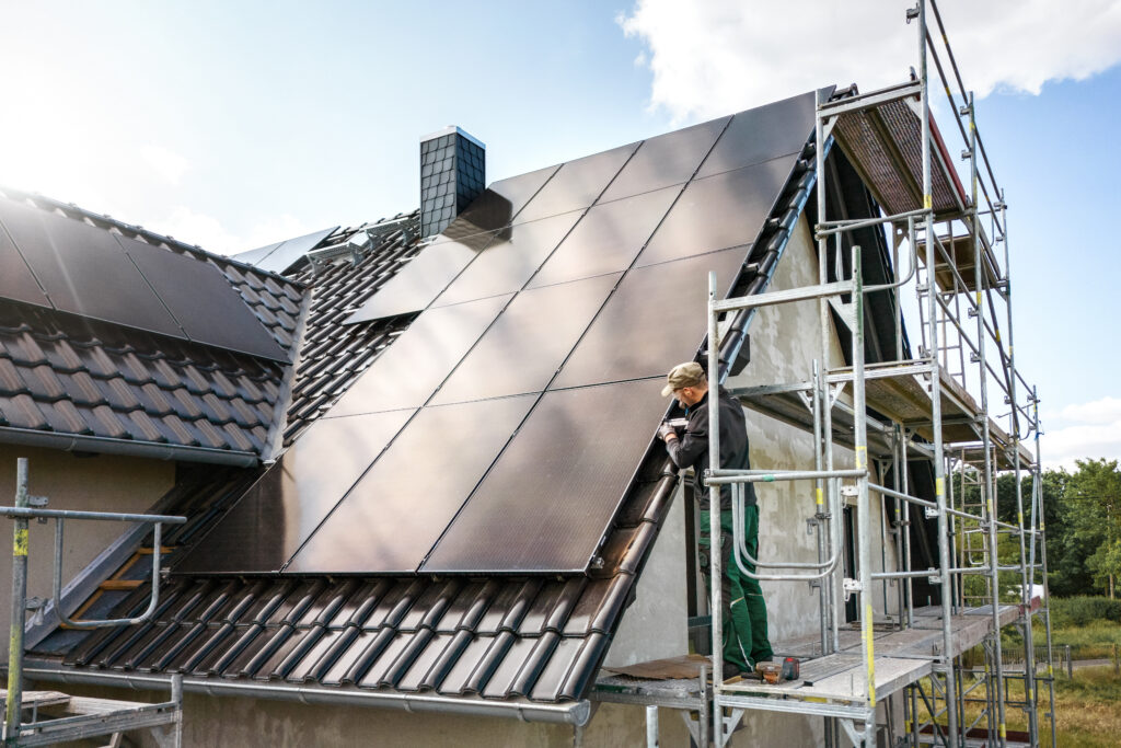 Ein Dach mit Solarpanelen zur Steigerung der Energie-Effizienz. An einer Seite des Daches ist ein Gerüst befestig. Darauf steht ein Mann, der an einem der Solarpanele arbeitet. 