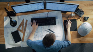 Symbolbild einer Person am Schreibtisch mit Monitor, Tastaur, Maus und einer Zeichnung, um den Alltag einer Energie-Effizienz-Experten darzustellen.